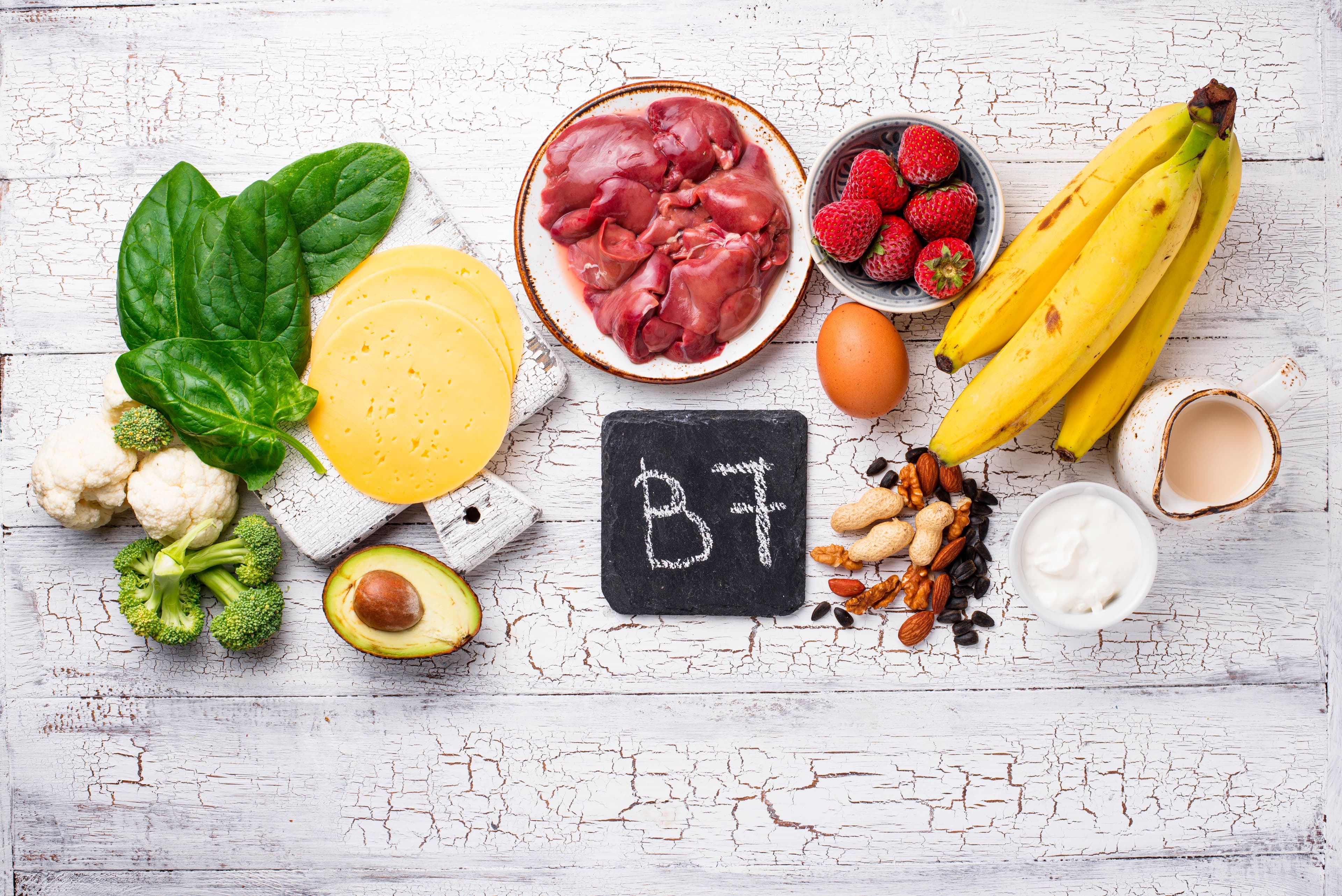 Foods rich in biotin (Vitamin B7) displayed on a rustic table, including bananas, eggs, nuts, cheese, liver, spinach, strawberries, avocado, and broccoli for healthy hair, skin, and nails.