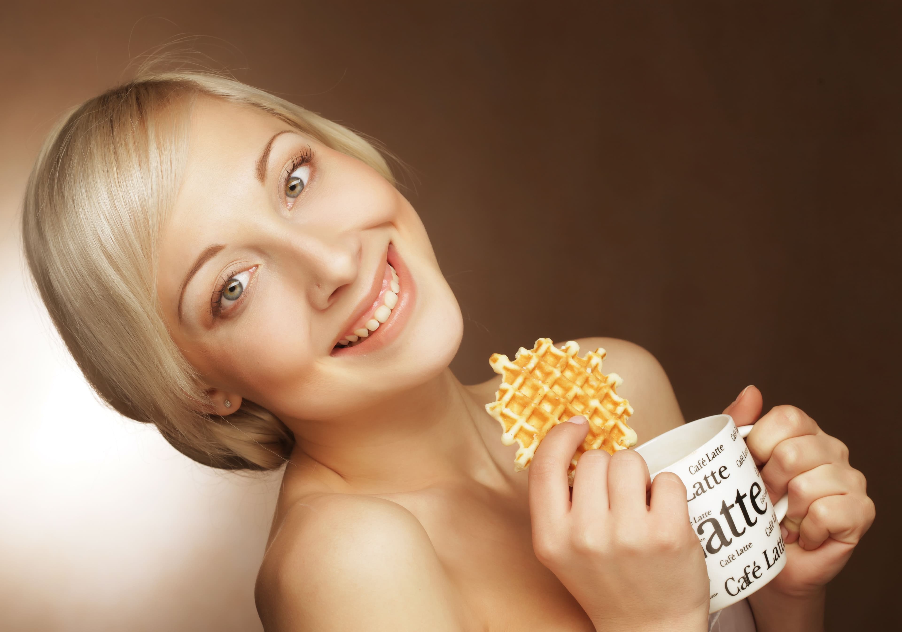 Smiling blonde woman holding a coffee mug and a waffle, symbolizing energy and vitality associated with glutathione benefits for health and wellness