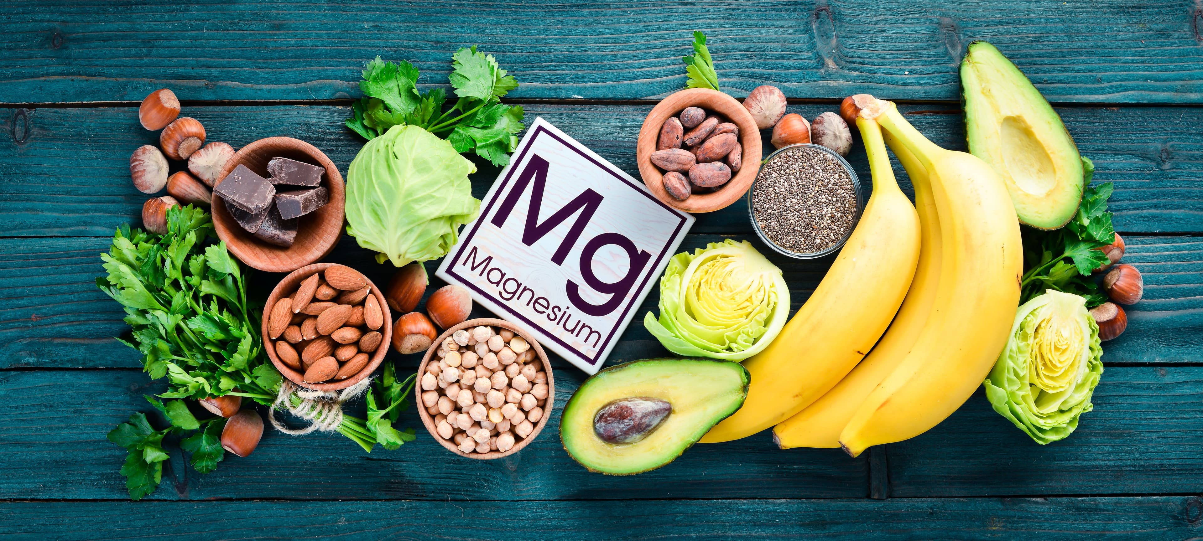 A selection of magnesium-rich foods, including bananas, avocados, almonds, dark chocolate, leafy greens, and seeds, displayed on a wooden table.